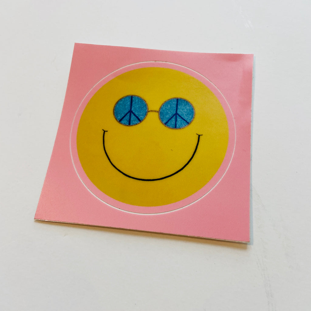 yellow smiley face with blue peace sign sunglasses on a pink background sticker